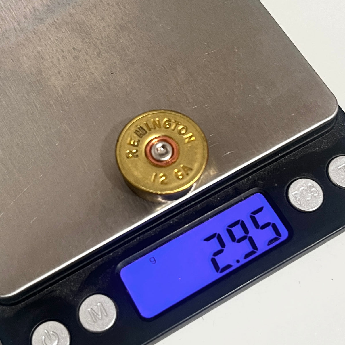 A brass Remington 12 Gauge gold headstamp sits on a digital scale displaying a weight of 2.95 grams with silver spent primer.