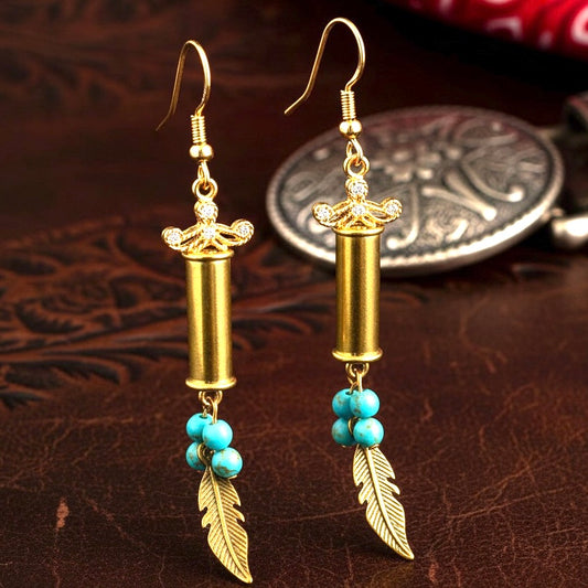 A pair of dangle earrings crafted from authentic, once-fired .22 caliber brass shells. The casings show visible fired wear and spent rim strikes. They are adorned with turquoise beads and golden feather charms, featuring a precision-drilled hole for the earring hardware.