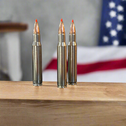3 nickel 30-06 dummy rifle rounds with red-tipped bullets standing on a wood surface in front of an American flag backdrop