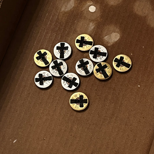 A collection of mixed gold and silver 12-gauge shotgun shell slices scattered on brown cardboard, featuring engraved black cross silhouettes on each authentic thin-cut piece.