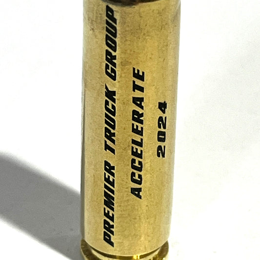 Close-up of a .50 BMG brass casing engraved with 'PREMIER TRUCK GROUP ACCELERATE 2024' text along its side