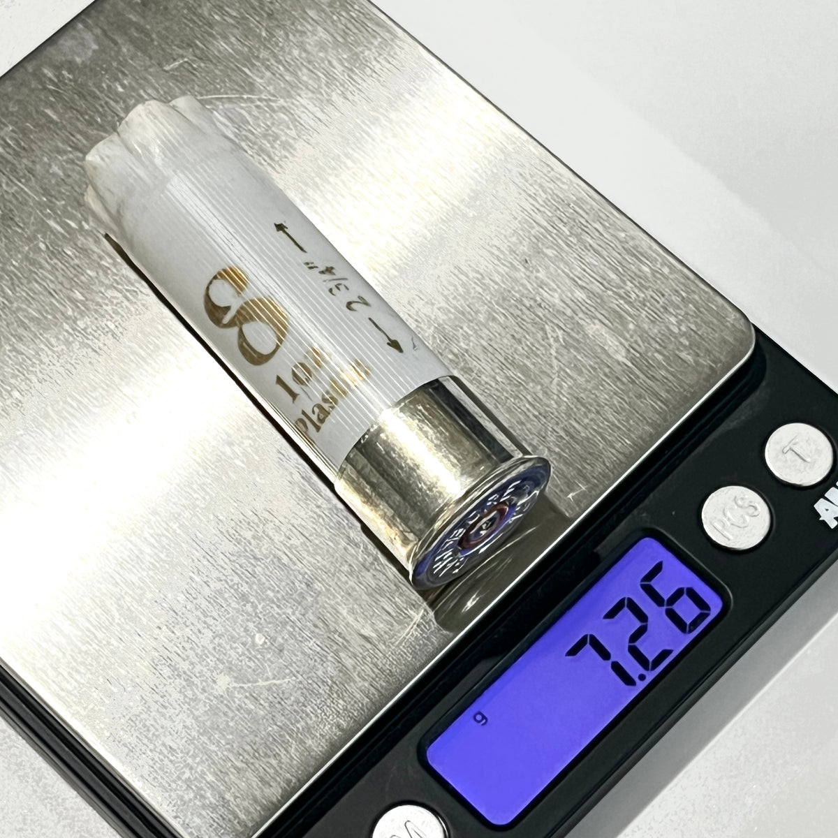 A single once fired white shotgun shells rests on a digital kitchen scale displaying 7.26 g.