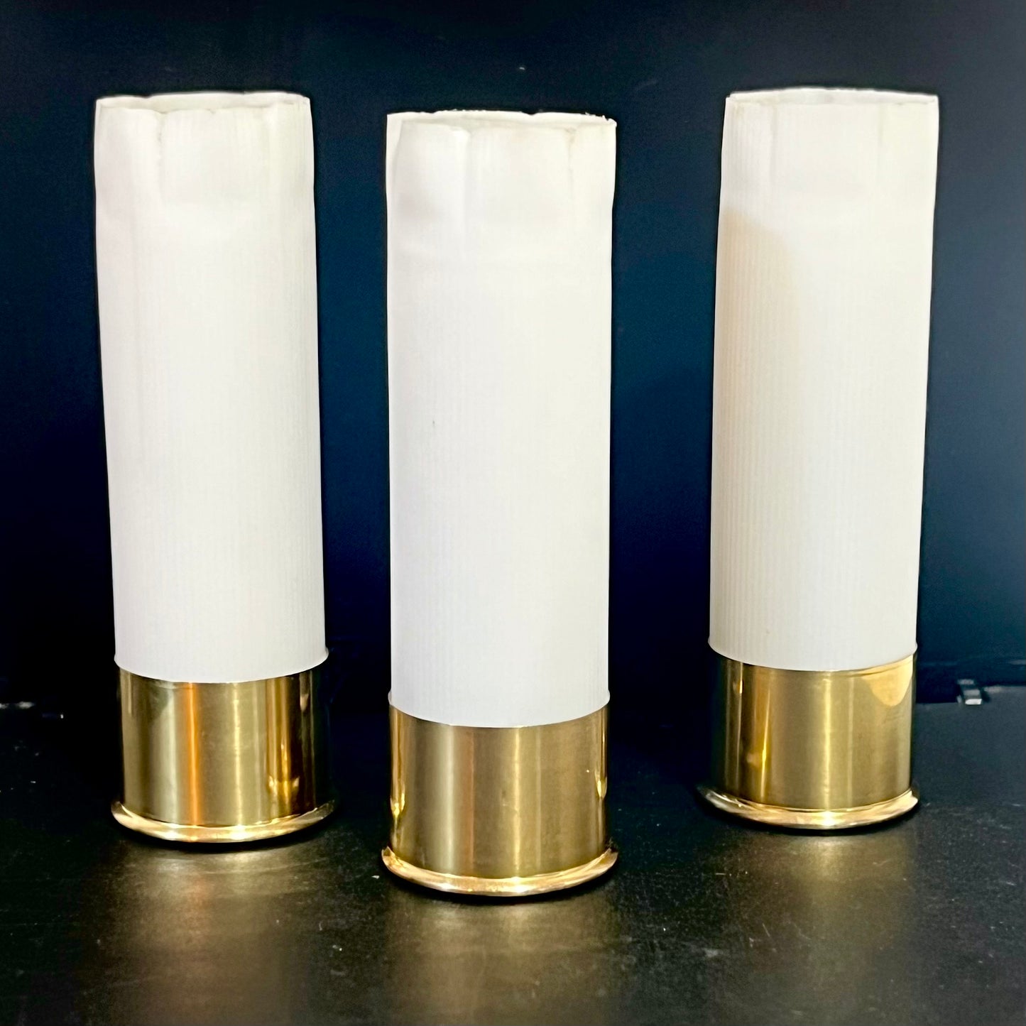 Three white 12 gauge blank shotgun shells with brass bases standing upright on a dark background
