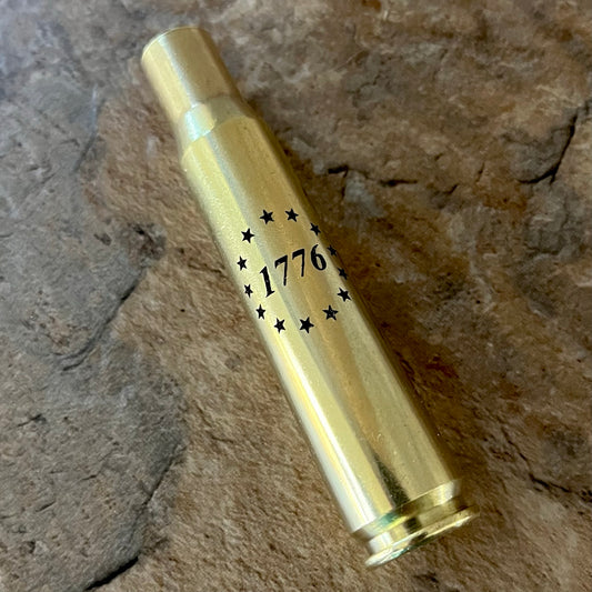 Used brass rifle shell engraved with '1776' and Betsy Ross 13 stars, resting on a natural stone surface.