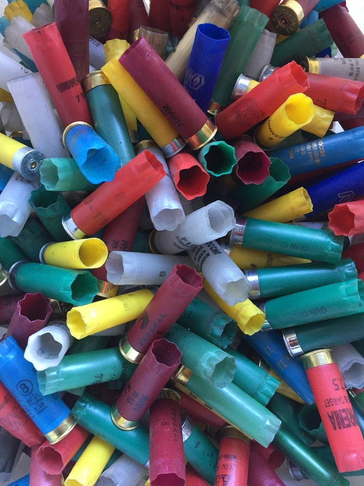 Scattered pile of recovered, once-fired 12-gauge empty shotgun shells in mixed colors (red, blue, green, pink). The ribbed plastic hulls feature silver and brass bases with open crimped ends for authentic DIY bulk crafting