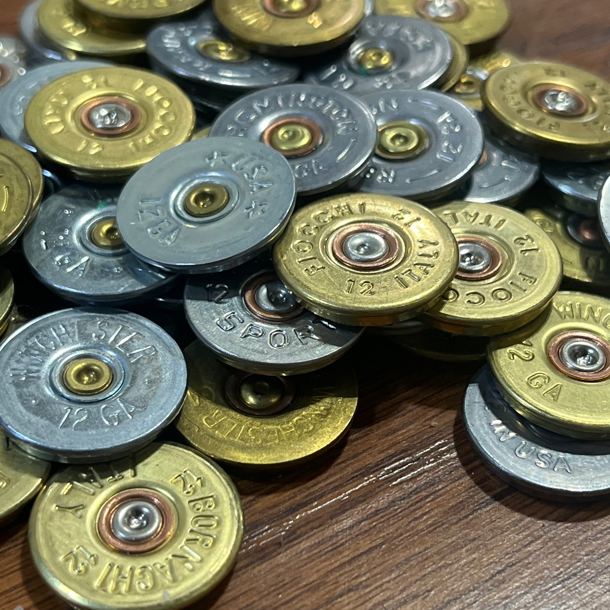 Close up bulk pile of mixed gold and silver 12 gauge shotgun shell headstamp slices on a wood background.