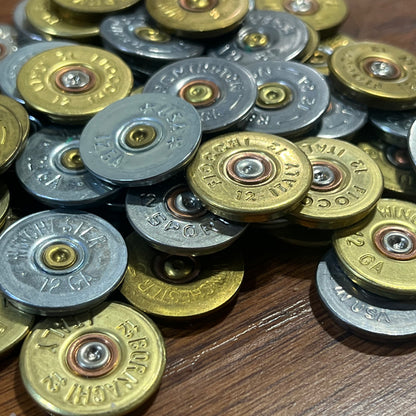 Close up bulk pile of mixed gold and silver 12 gauge shotgun shell headstamp slices on a wood background.