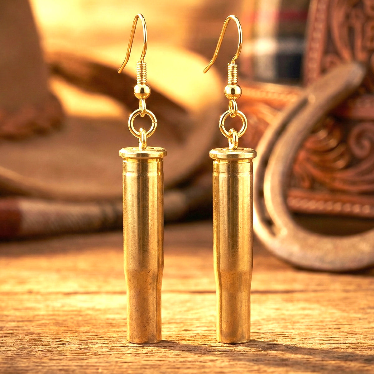 Pair of gold-tone .22 Hornet brass shell casings made into dangle earrings, displayed on a wood surface with a horseshoe and boots in the background