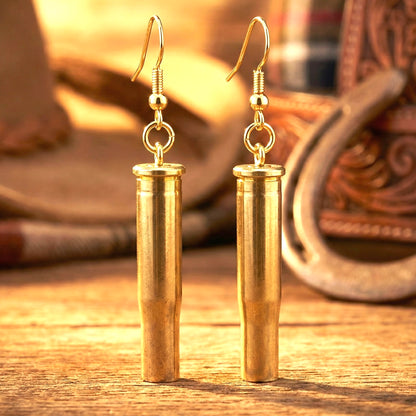 Pair of gold-tone .22 Hornet brass shell casings made into dangle earrings, displayed on a wood surface with a horseshoe and boots in the background