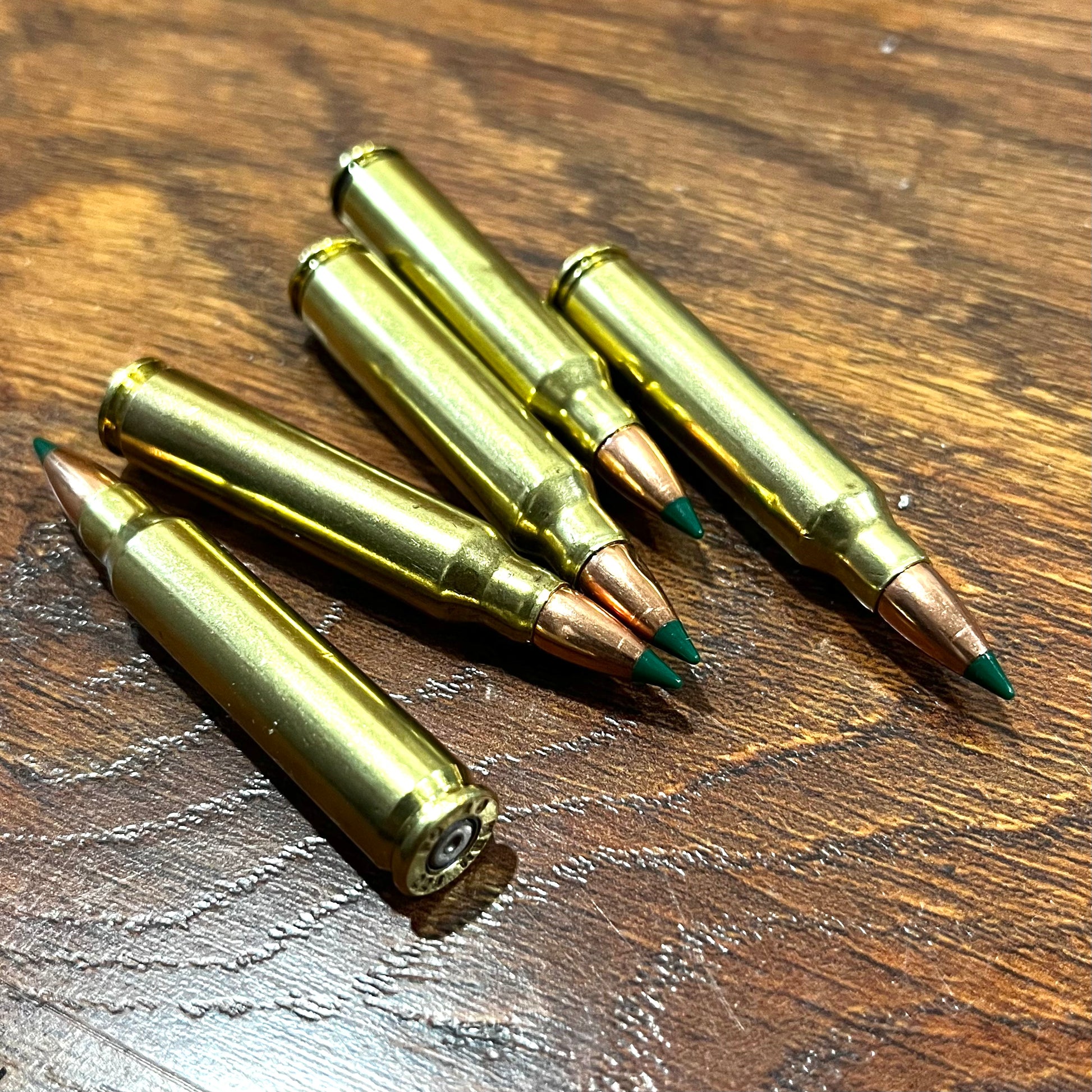 Five inert 5.56 brass training rounds with green tips grouped diagonally on a dark textured wood surface.