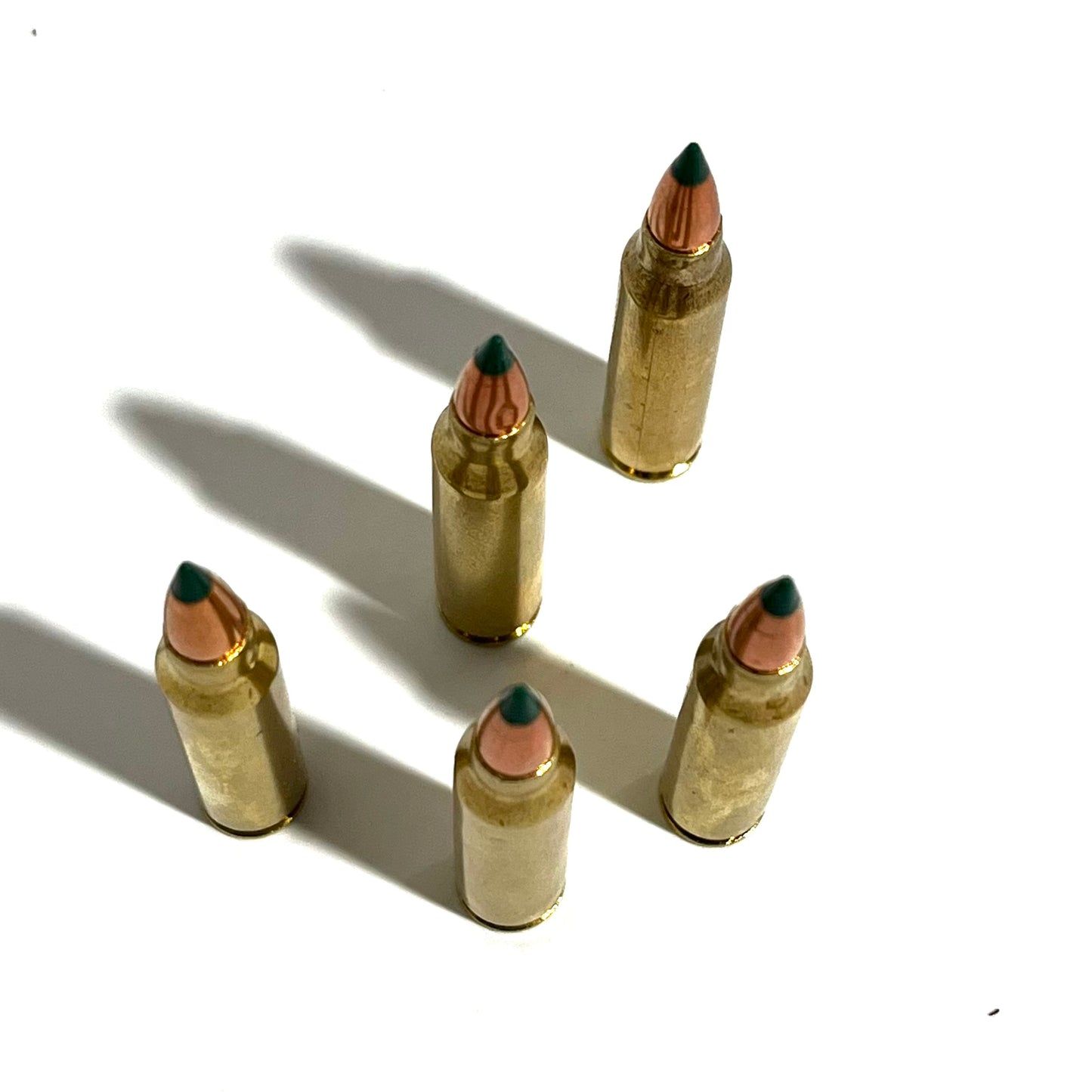 Five brass 5.56 replica cartridges with green tips standing upright on a white surface, casting strong shadows.