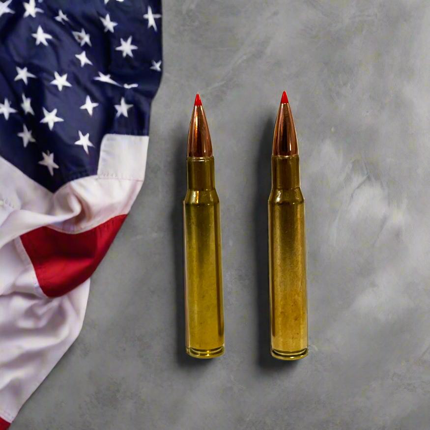 Two 30-06 brass red tip fake casings standing on a gray surface beside an American flag.