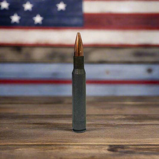 Steel 30-06 dummy rifle round standing upright on a wood surface in front of an American flag backdrop