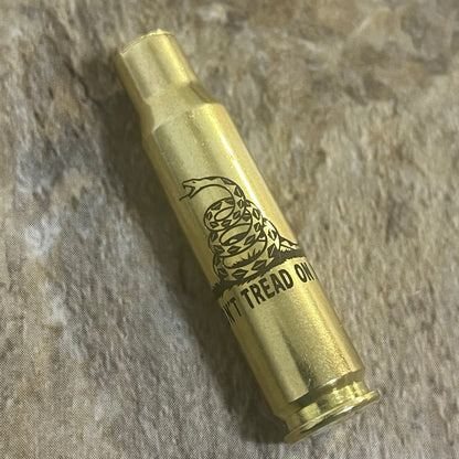 Single 308 WIN brass shell with laser engraved coiled Gadsden rattlesnake lying diagonally on a stone surface.