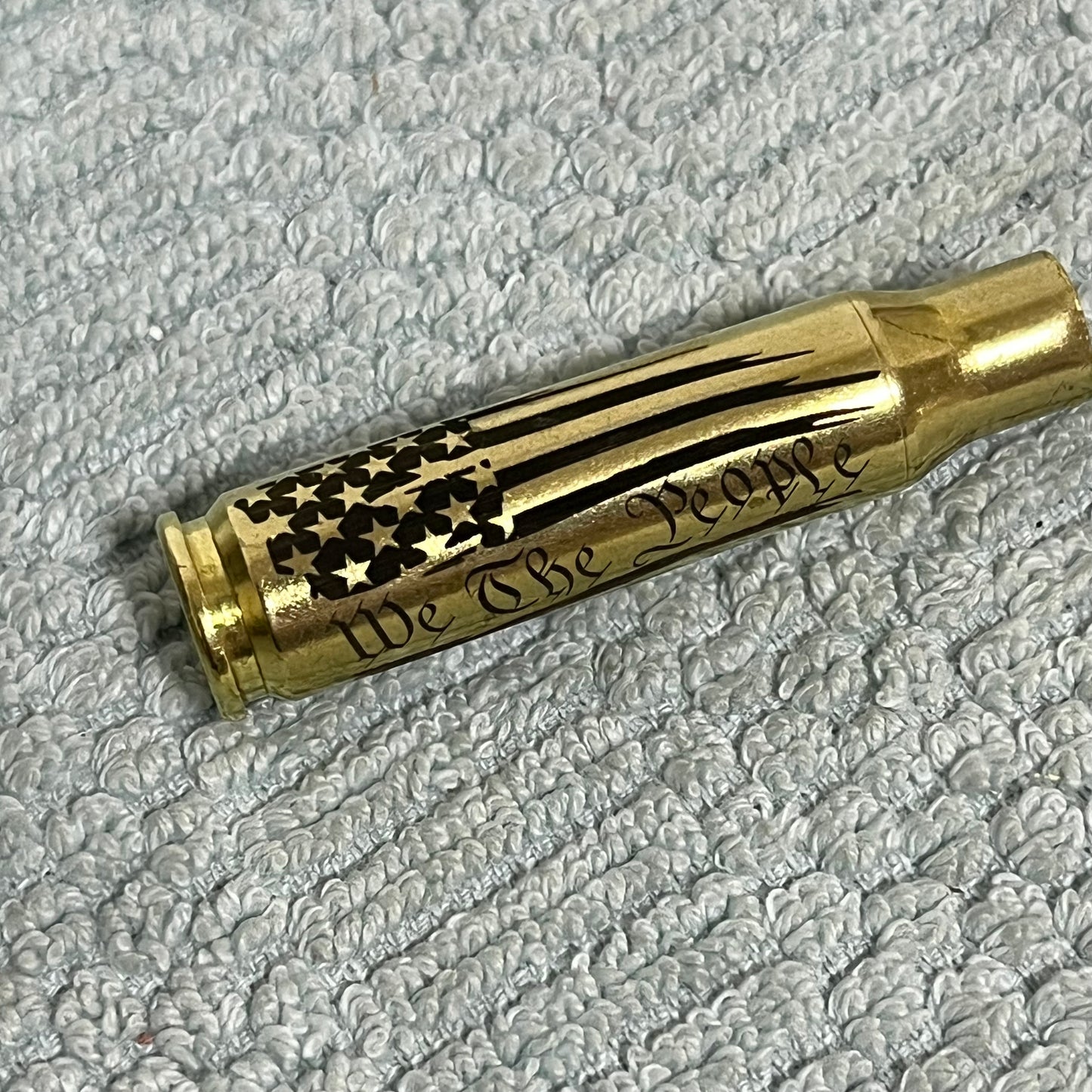 One horizontal .308 WIN cartridge case engraved with the We The People flag design on a white textured fabric surface.
