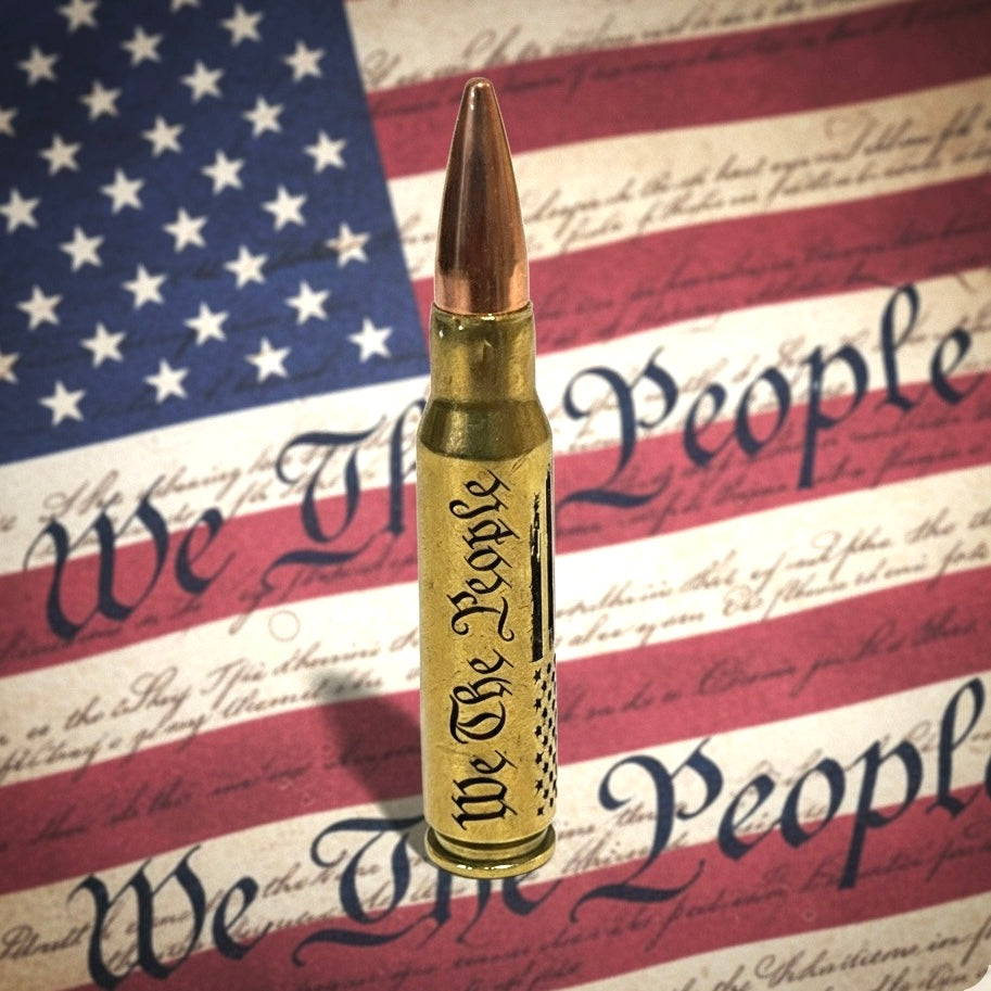 308 win dummy round with We The People and flag engraving on authentic once-fired brass against a flag backdrop.
