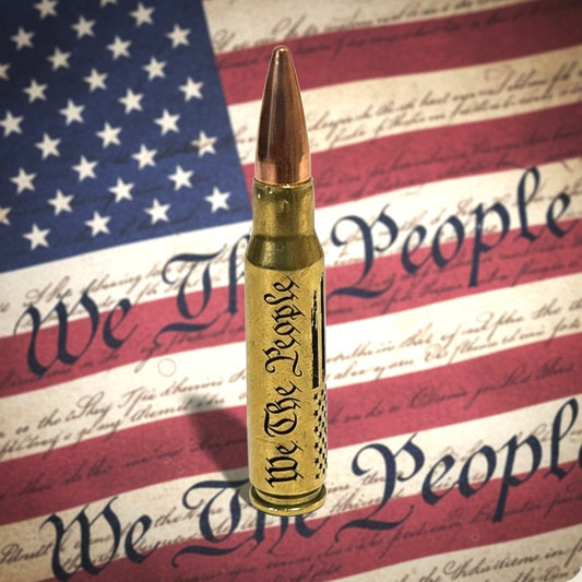 308 win dummy round with We The People and flag engraving on authentic once-fired brass against a flag backdrop.
