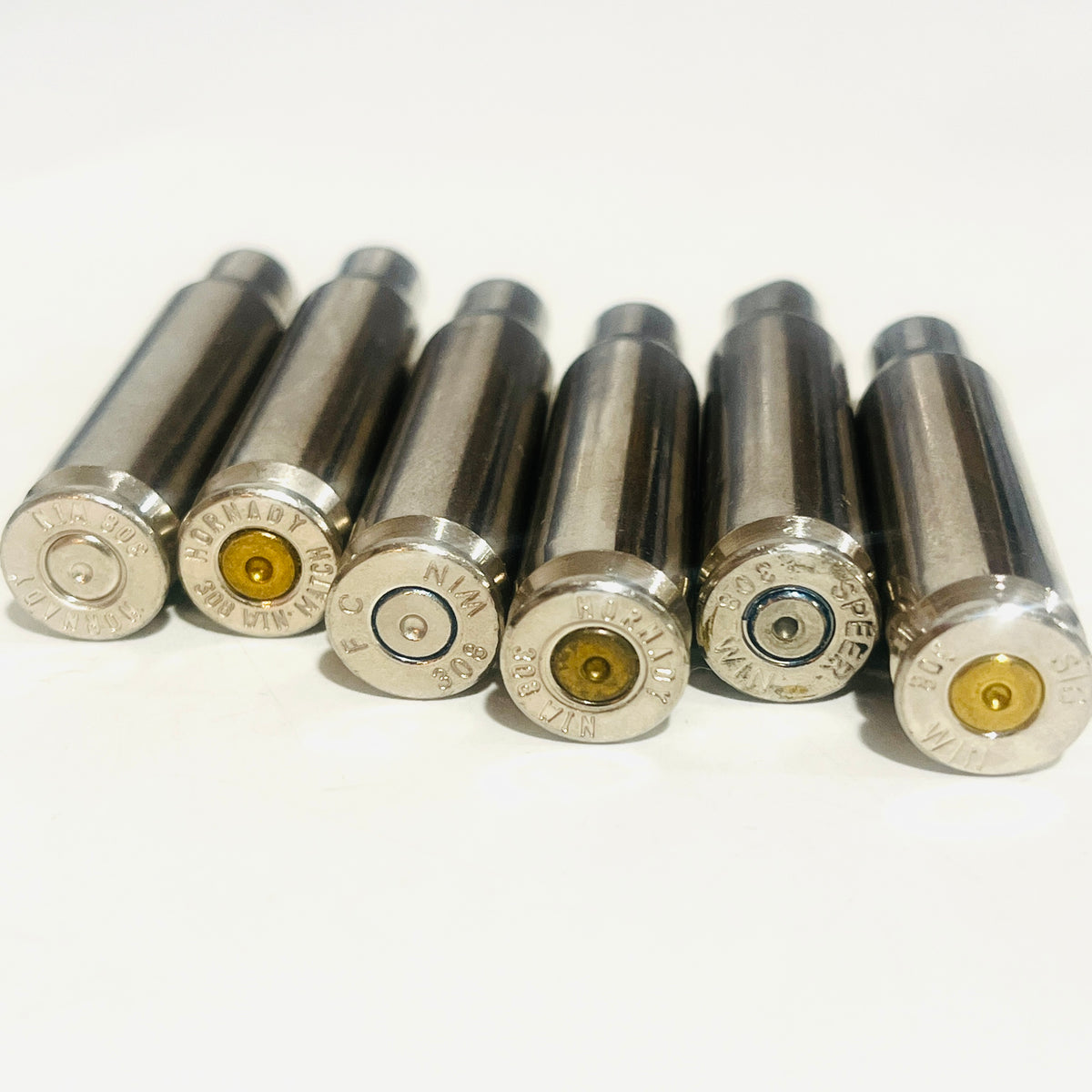 Six empty silver-tone .308 WIN nickel-plated brass casings resting on a white surface, with bases and silver and gold spent primers facing the camera.