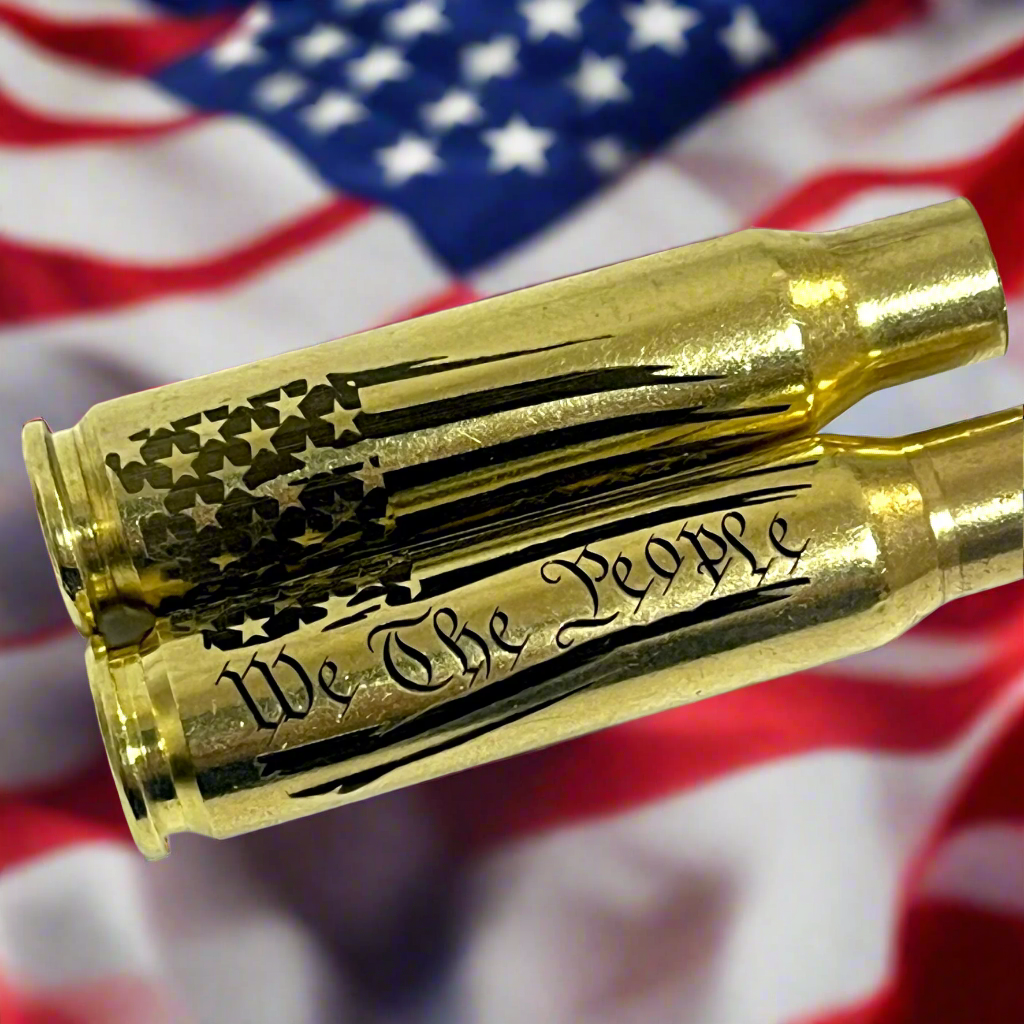 Two .308 WIN spent casings engraved with We The People and flag patterns, staged against a blurred American flag.