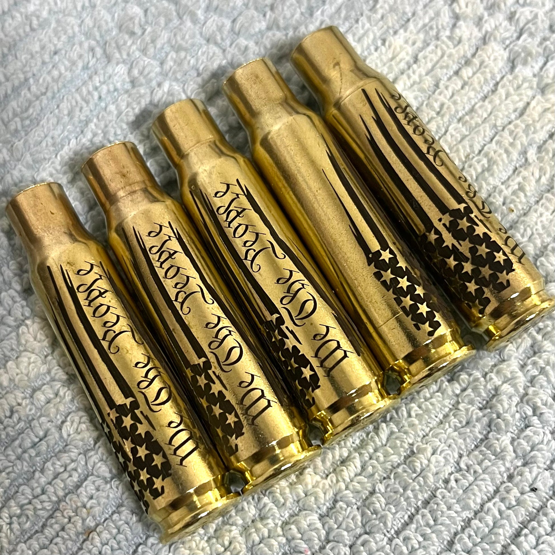 Five horizontal .308 WIN spent casings engraved with a We The People flag design, lined up on a white textured surface.