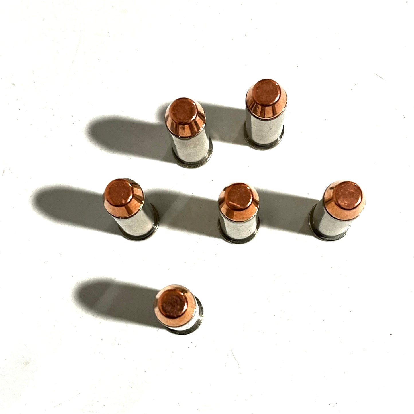 Six nickel display fake cartridges with copper bullets standing upright on a white surface, casting long dark shadows.