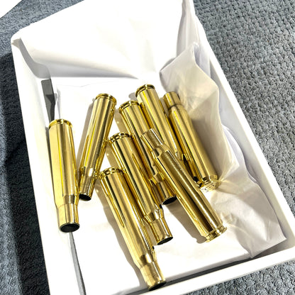 A set of shiny gold hand  polishd 50 cal spent cases arranged inside an open white box lined with white tissue paper, photographed from above on a gray textured surface.