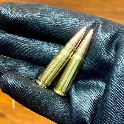 Two 7.62x39 dummy rounds with copper bullets held in a hand wearing a black textured glove.
