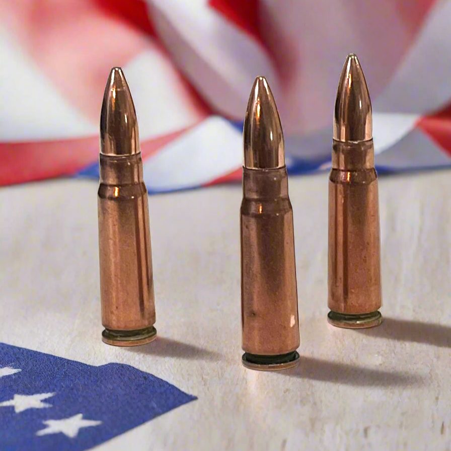 3 copper 7.62x39 dummy rounds standing upright on a wood table in front of a blurred American flag