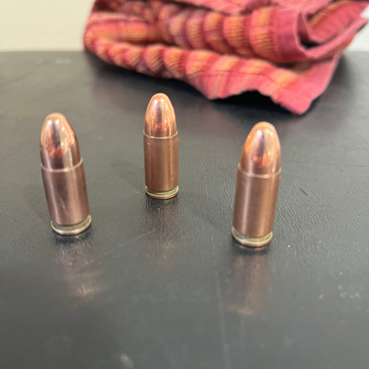 3 copper 9mm Luger dummy rounds standing upright on a dark surface with a striped cloth in the background
