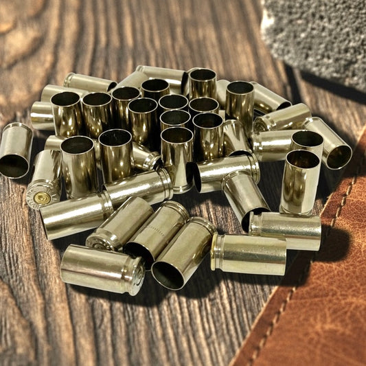 Small pile of nickel 9mm empty casings on a wood-grain surface next to a brown leather item, one casing showing a visible headstamp