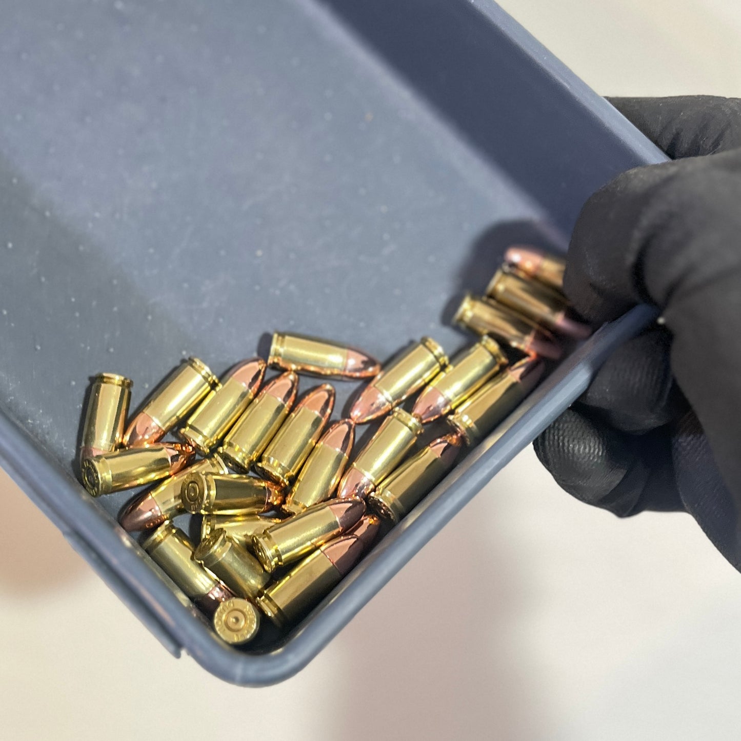 A hand wearing a black glove holds a gray plastic tray containing several brass 9mm dummy rounds and copper-colored bullets.
