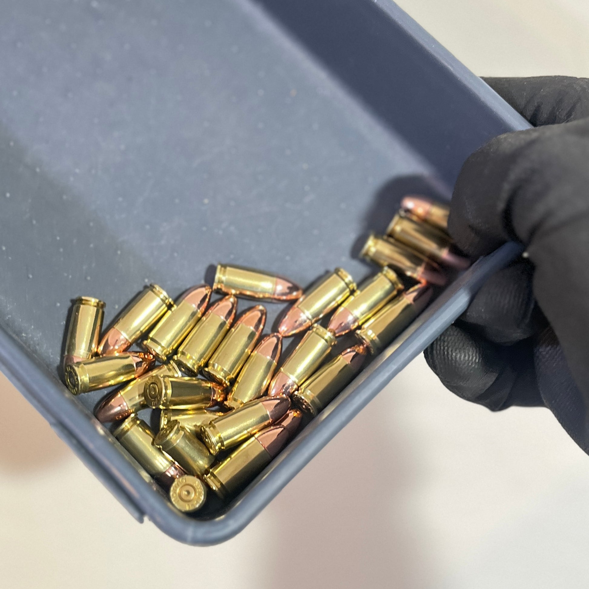 A hand wearing a black glove holds a gray plastic tray containing several brass 9mm dummy rounds and copper-colored bullets.