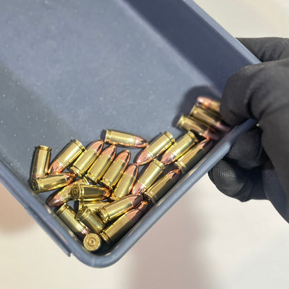 A hand wearing a black glove holds a gray plastic tray containing several brass 9mm dummy rounds and copper-colored bullets.