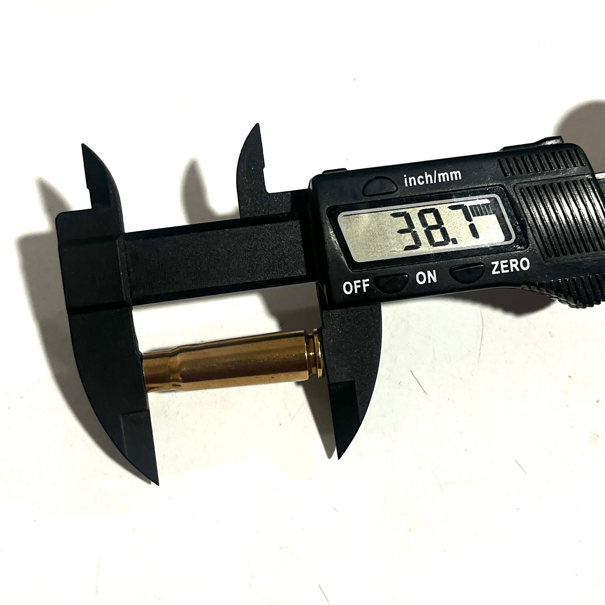 Empty brass casing measured lengthwise with a digital caliper, showing a reading of 38.7 mm on a tan background.