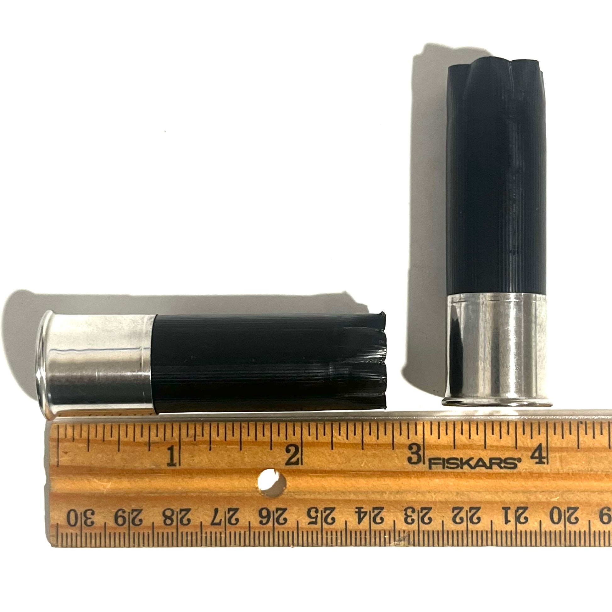 Two black shotgun shells with high brass bottoms beside a ruler showing a length of about 2.75 inches