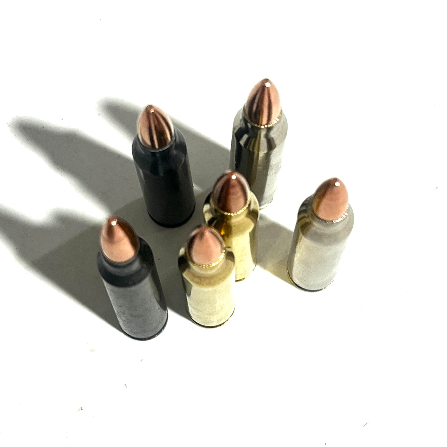 Six .223/5.56 shells in black, brass, and silver finishes stand together at an angle, casting shadows.