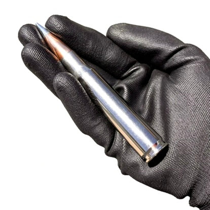 Black textured gloved hand holding a silver nickel-plated 50 cal BMG dummy round with a sharp copper bullet tip.
