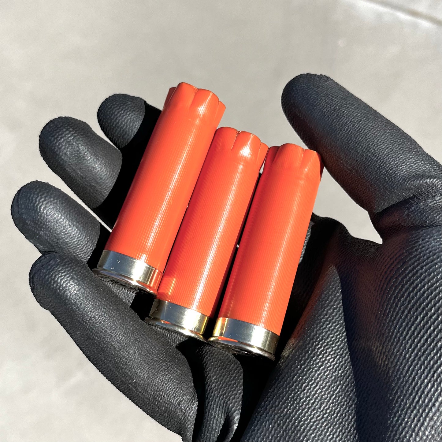 Gloved hand holding three blank orange 12 gauge shotgun shells with gold metallic bases.
