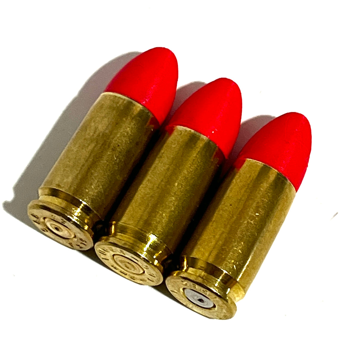 3 fake 9mm Luger dummy rounds with red painted tips laid diagonally on white background, brass casings visible