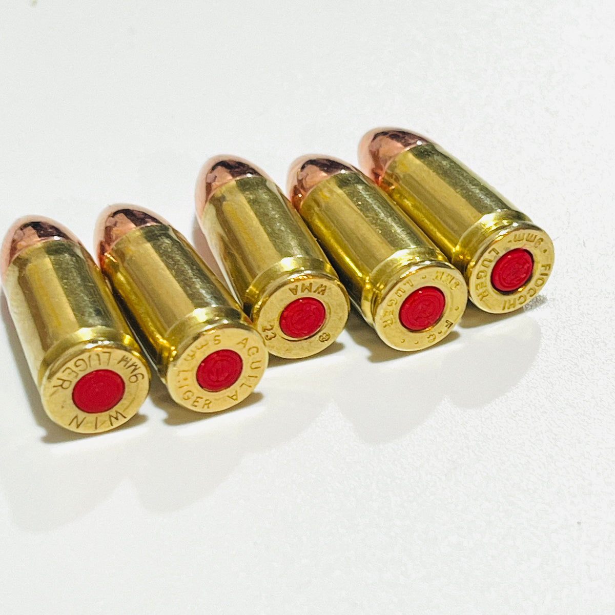Five used brass 9mm Luger cartridges in a row showing WIN, Aguila, WMA, FC and Fiocchi headstamps with red primers