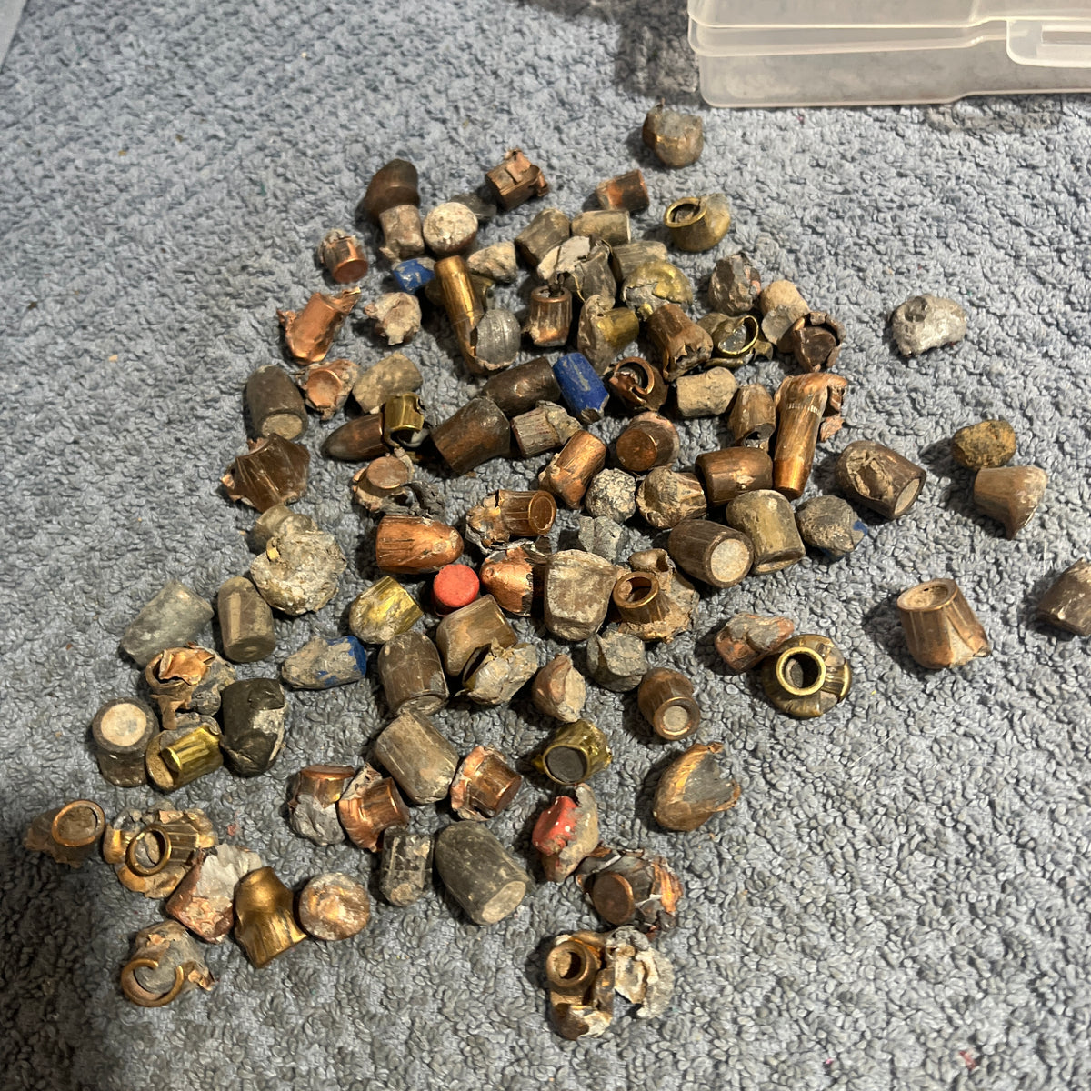 Pile of deformed spent bullet fragments on a gray towel next to a plastic ammo case