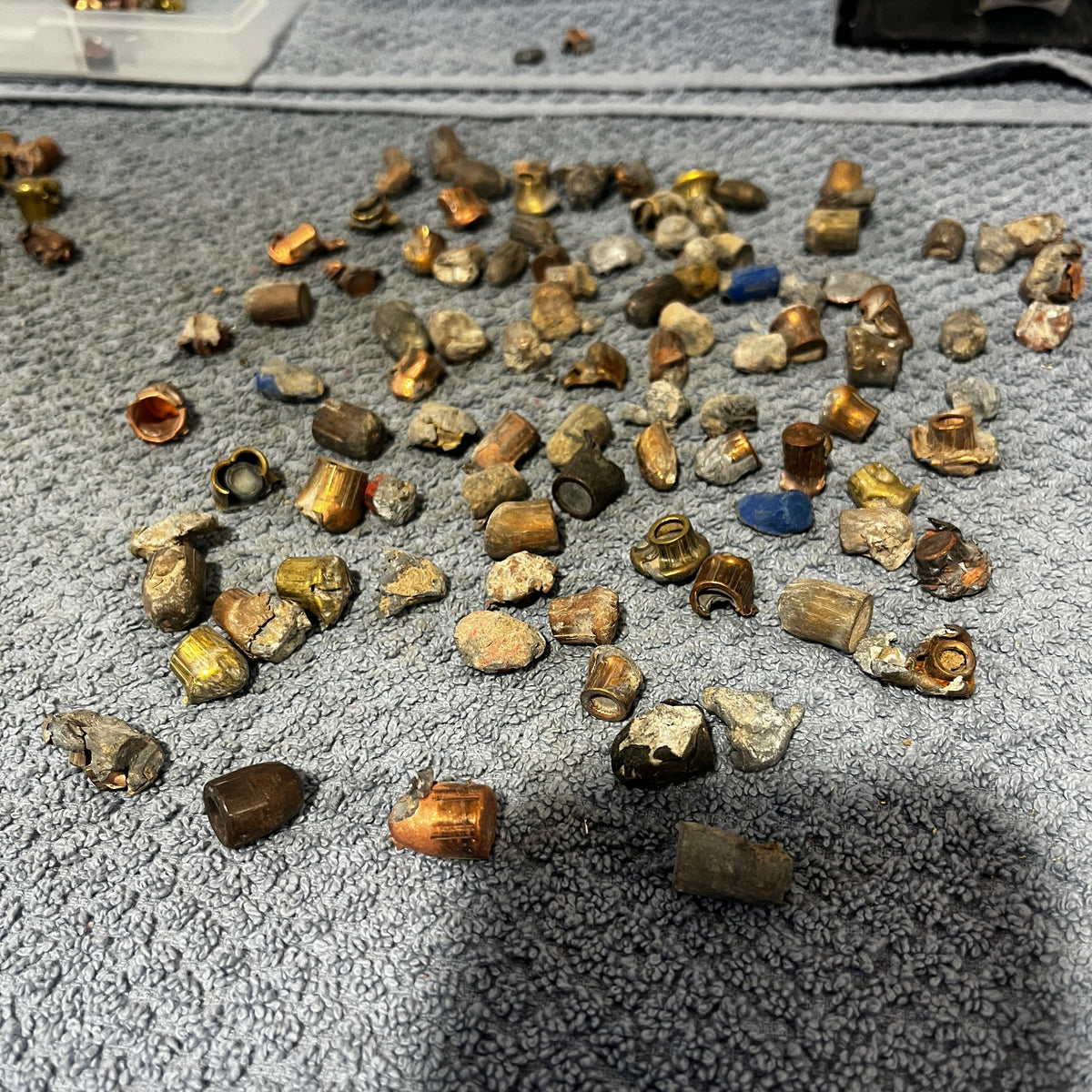Pile of deformed spent bullet fragments on a gray towel next to a plastic storage box