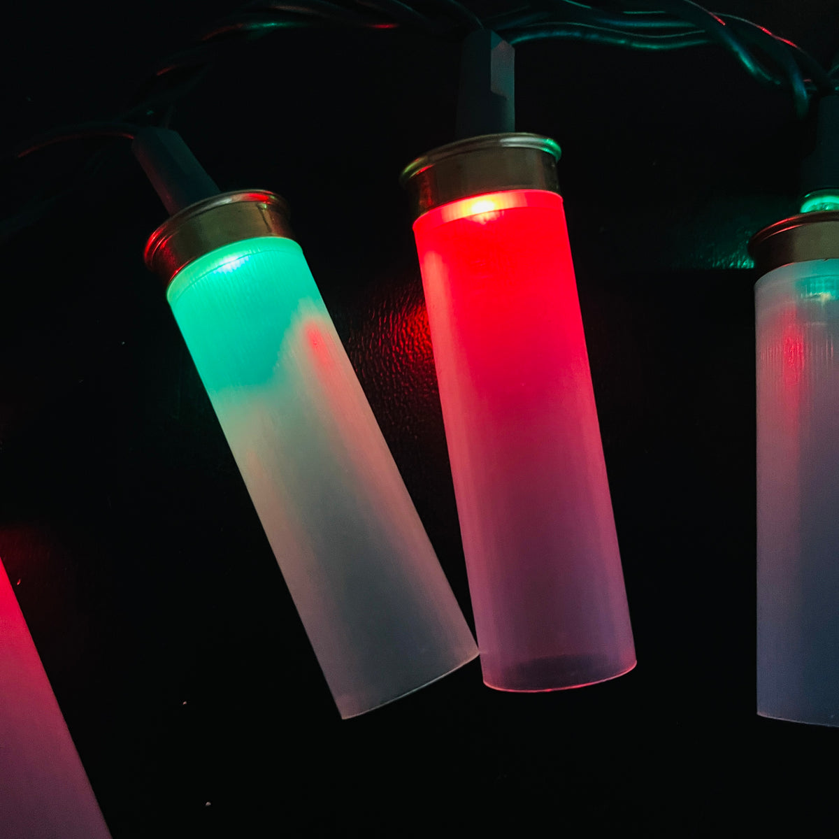Close-up of two clear empty shotshells hanging from a light strand, one glowing green and one glowing red in the dark