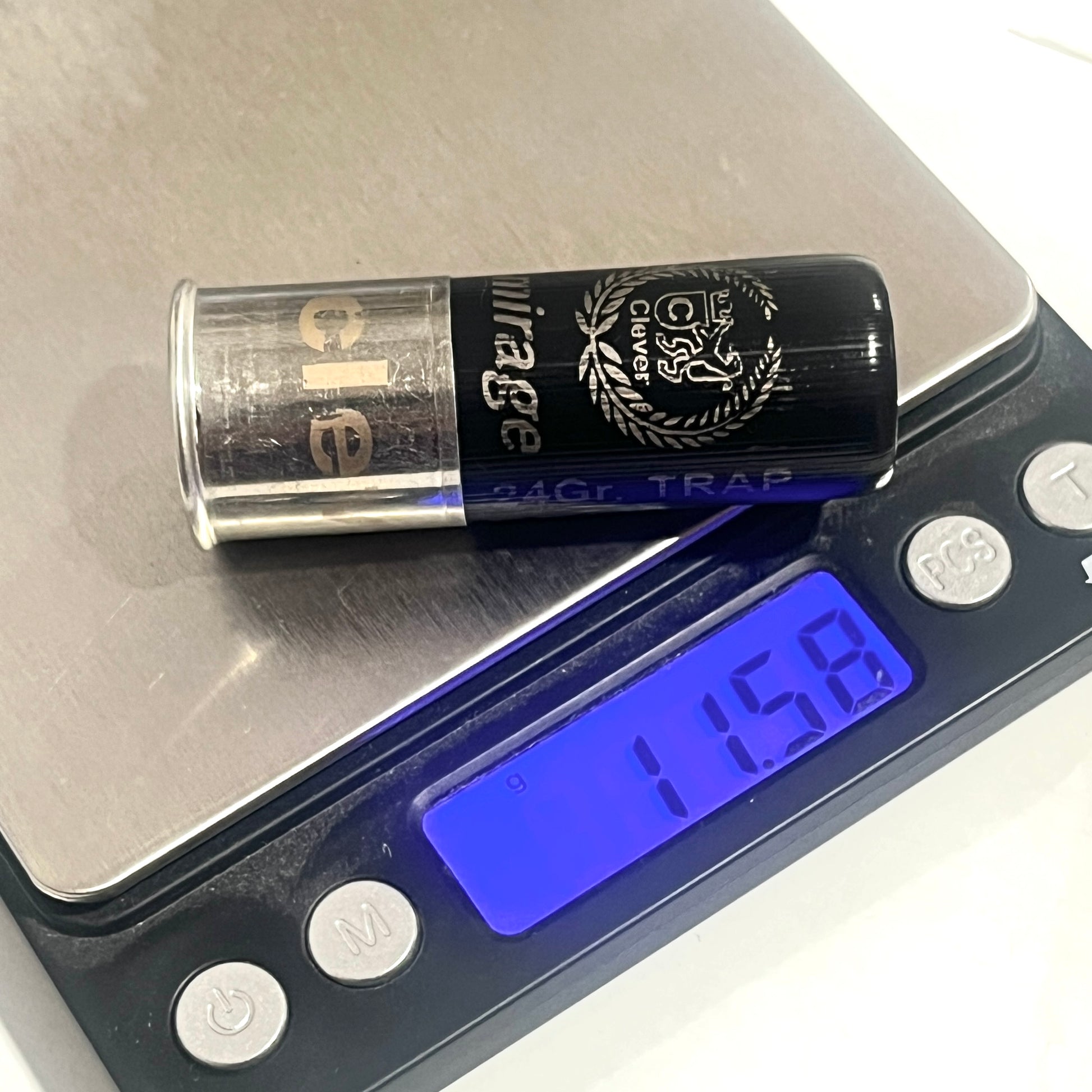 Digital scale weighing a black 12-gauge shotshell, marked "24Gr. TRAP" and "mirage," reading 11.58.