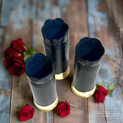 Three dark blue 12 gauge blank shotgun shells with gold bases arranged among red roses on a rustic wood surface