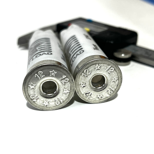 Close-up of two deprimed white Stars and Stripes 12 gauge shotgun shell bases showing empty primer holes next to a caliper