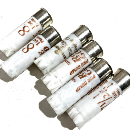 A row of six white 12 gauge empty shotgun shells lined up diagonally on a white background, highlighting the silver bases and printed load details.
