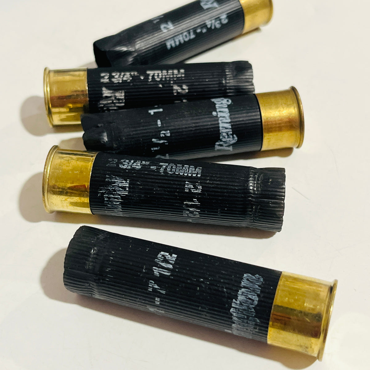 Five empty black shotgun hulls with brass bases scattered on a white surface, printed 2 3/4" - 70MM