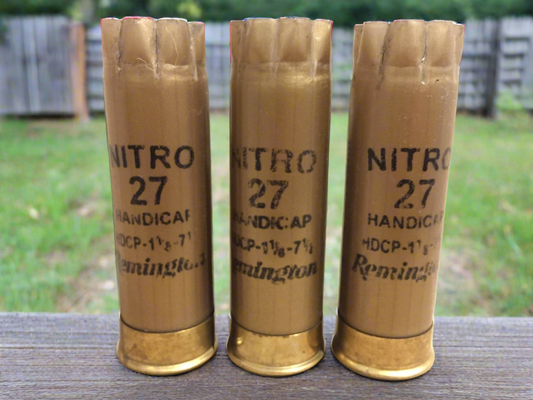 Three gold empty Remington Nitro 27 shells standing on a wooden surface.
