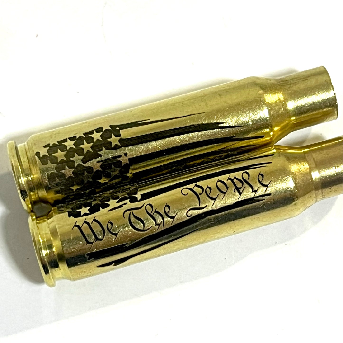 Two horizontal .308 WIN brass casings engraved with the We The People flag pattern on a solid white surface.